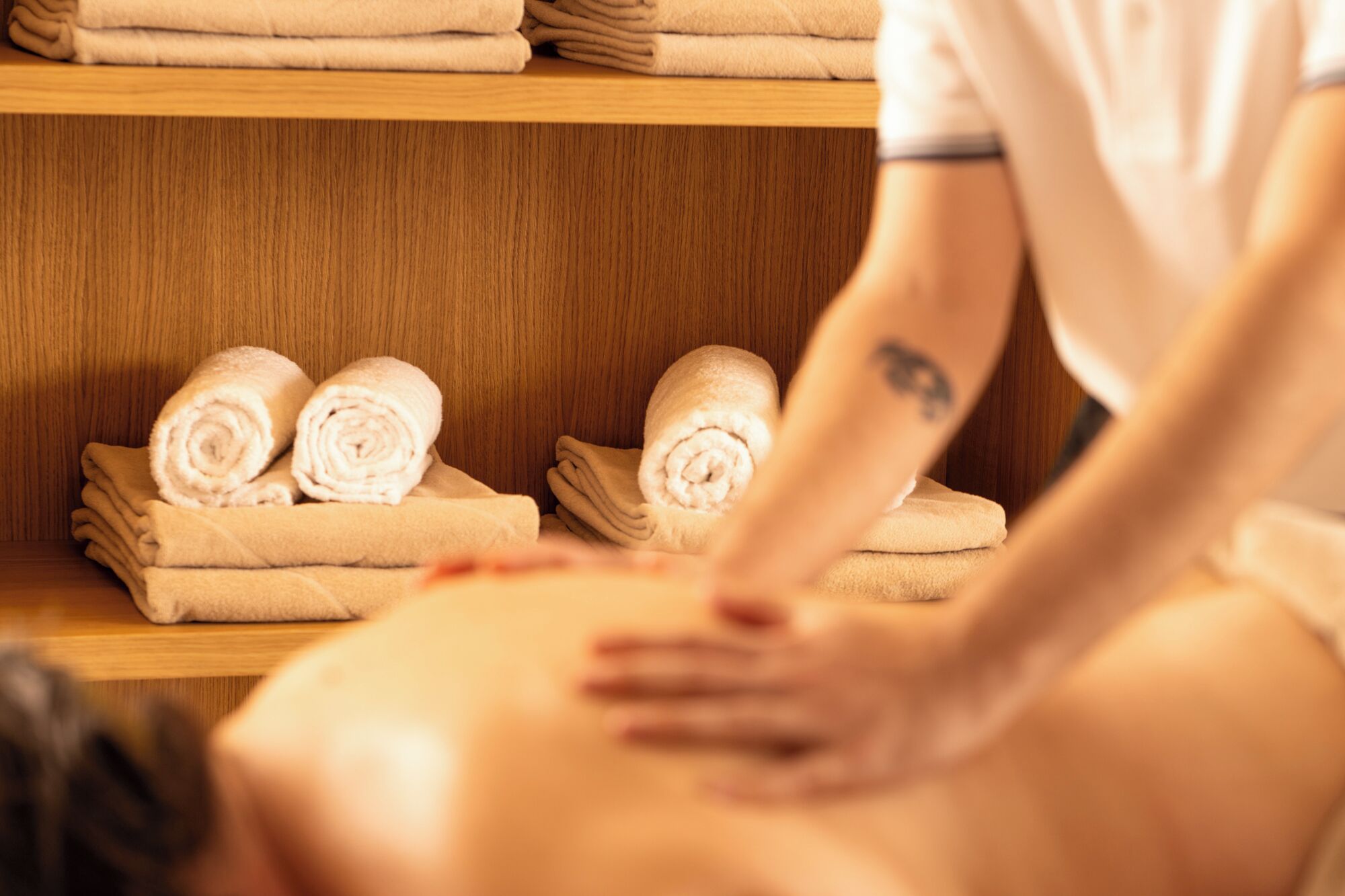 A person receiving a back massage in a room with wooden shelves holding neatly folded towels.