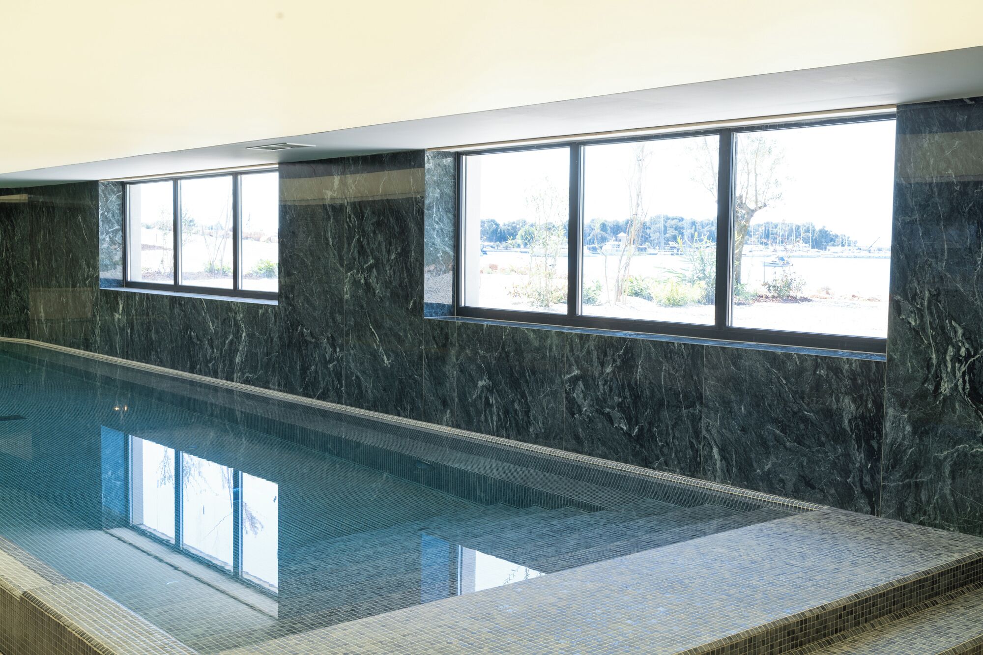 A swimming pool with a tiled edge, large windows, and a view of trees and buildings.