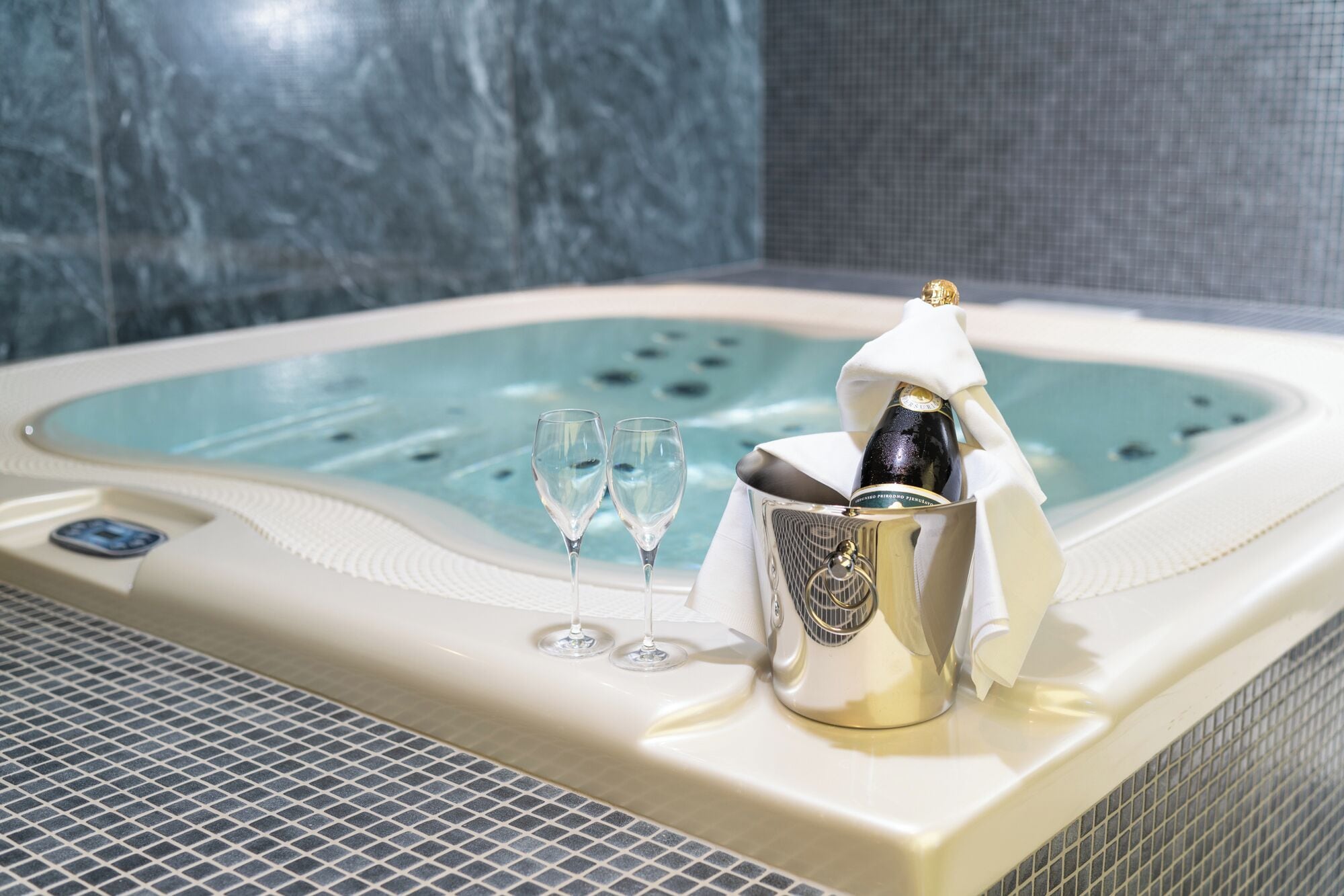 A hot tub with champagne and two glasses, a control panel, and a tiled floor.