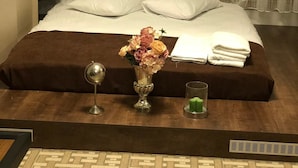 Una cama bien hecha con almohadas blancas y una colcha marrón, un florero con flores, un espejo redondo pequeño y un vaso de agua sobre una mesita de noche de madera.