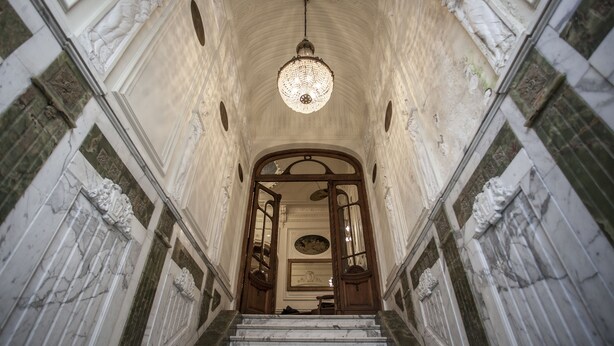 Un magnifique escalier en marbre, avec des marches de marbre et des portes en bois richement décorées, menant à un plafond haut orné d’un lustre en cristal.