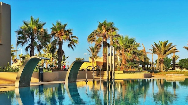 Une piscine aux eaux cristallines, au design moderne, entourée de palmiers et surplombée d’un ciel d’un bleu éclatant.
