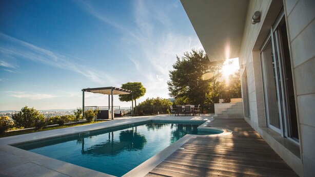 Zona de piscina moderna al aire libre con terraza de madera y pérgola, rodeada de vegetación y vistas a la ciudad.
