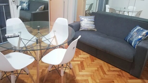 Una sala de estar moderna con una mesa de vidrio, sillas blancas, un sofá gris y piso de madera con patrón de espiga.