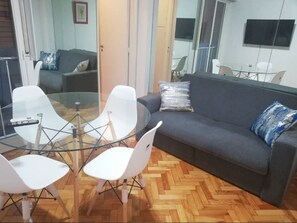A modern living room with a glass table, white chairs, a grey sofa, and a herringbone wooden floor. - Arenales & Austria (Buenos Aires)