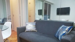 Un salón con un sofá gris, una mesa de vidrio y una silla blanca. Hay un televisor montado en la pared y una puerta con el letrero número 11.