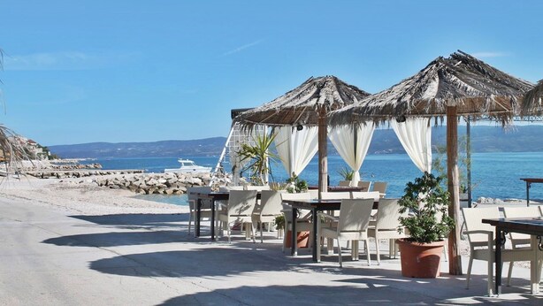 Un área de estar al aire libre con mesas y sillas, sombrillas de paja y plantas en macetas, con vistas a una playa y al mar.