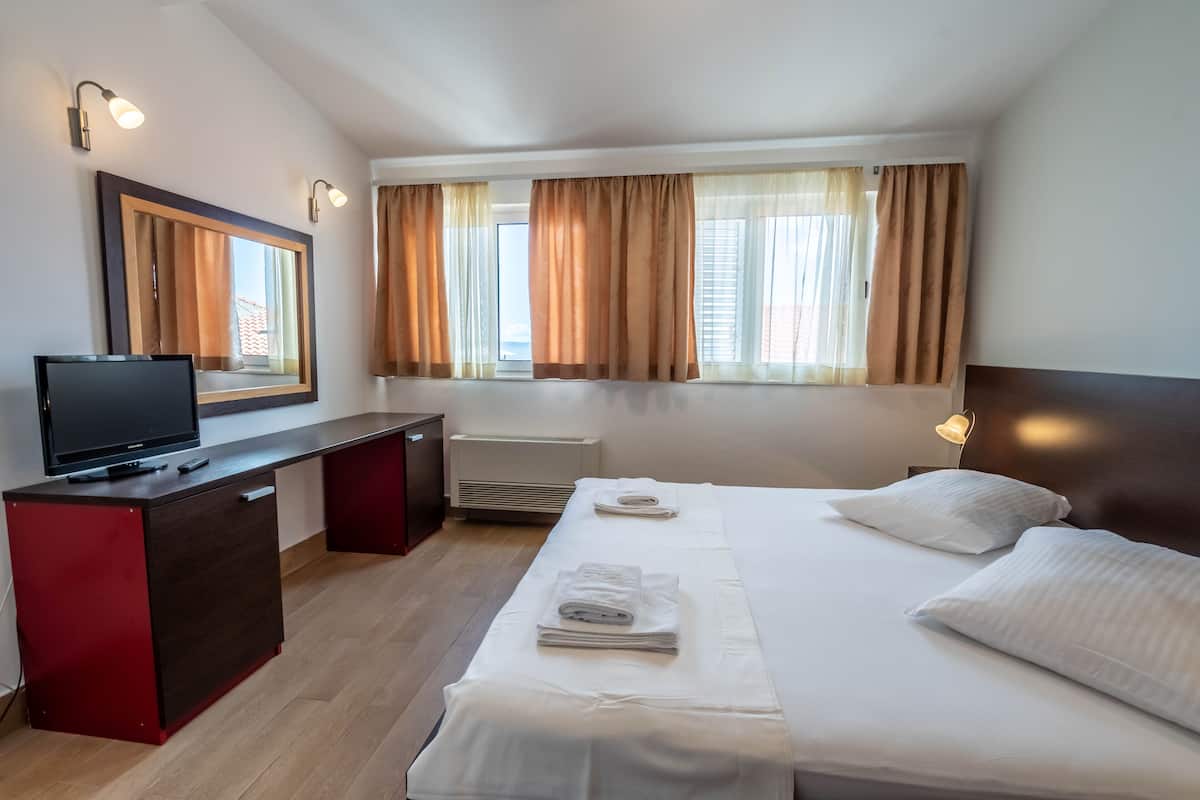 A hotel room with a bed, a desk with a TV, a mirror, and a window with curtains.