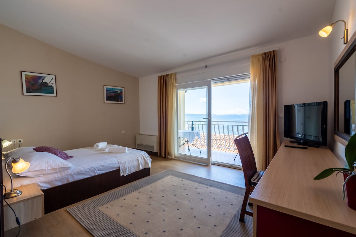A hotel room with two beds, a TV, a desk, and a balcony with a view of the sea.