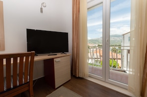 A room with a TV on a wooden desk, a chair, a sliding glass door to a balcony with a view of buildings and mountains. - Pansion Epetium (Split)