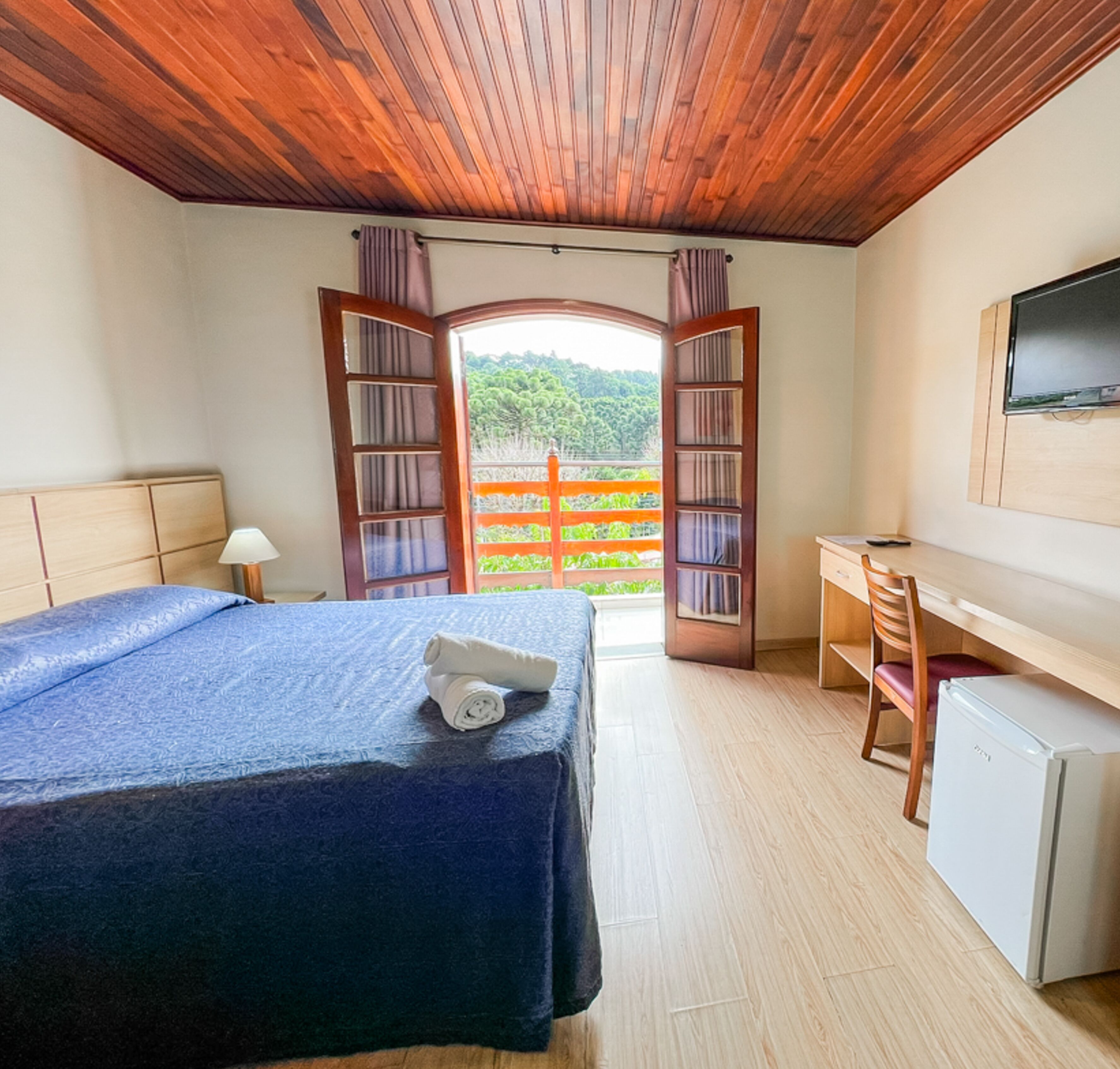 Un dormitorio con cama, escritorio, silla, televisor y balcón con vistas a zonas verdes.