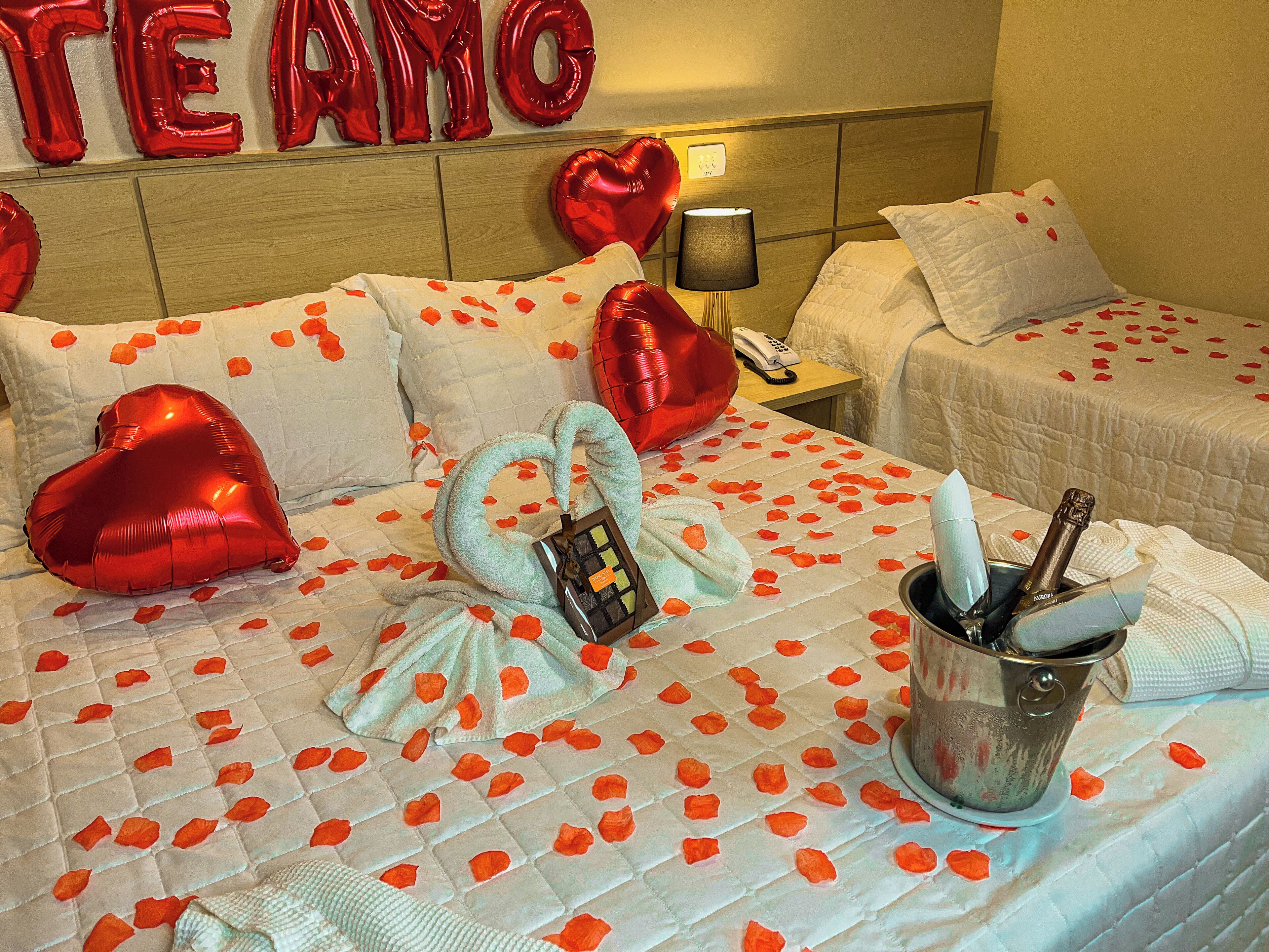 Ein Hotelzimmer mit einem Bett, das mit Rosenblüten, herzförmigen Ballons und einer Champagnerflasche dekoriert ist.