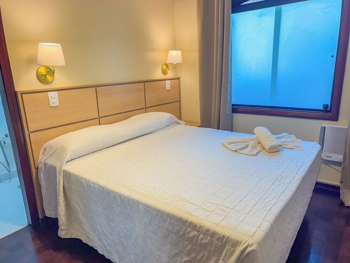 A hotel room with a bed, two wall-mounted lamps, a window with curtains, and a radiator.