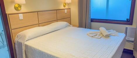 A hotel room with a bed, two wall-mounted lamps, a window with curtains, and a radiator.
