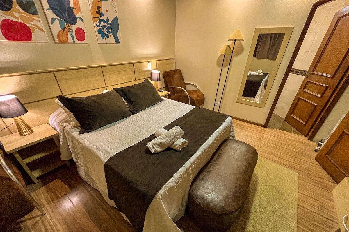 Habitación de hotel con cama, mesita de noche, silla, lámpara de pie, espejo y armario.