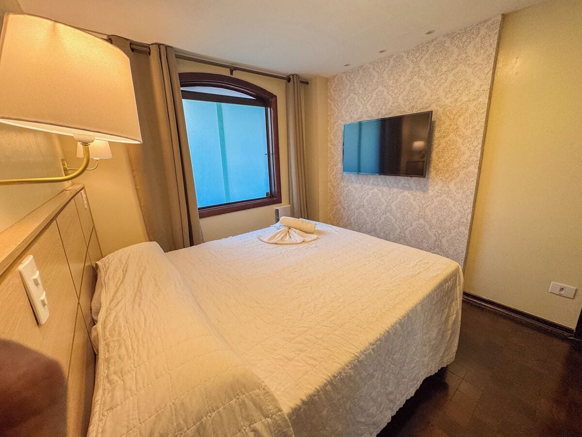 A hotel room with a bed, a TV mounted on the wall, a window with curtains, and a bedside lamp.