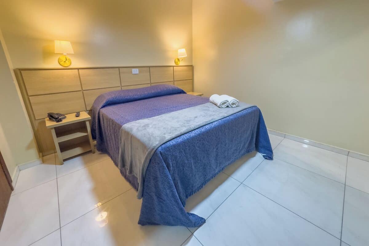 Una cama bien hecha con una colcha azul y gris, una mesita de noche con lámpara y teléfono.