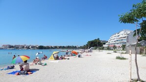 Ein Strand mit Personen, die sonnenbaden und schwimmen, Sonnenschirmen und einem Gebäude im Hintergrund.