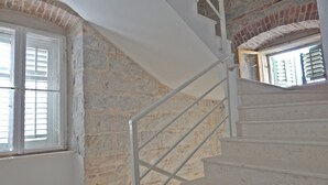 Una escalera con barandillas blancas que conduce a un rellano, rodeado de muros de piedra y una ventana con contraventanas.