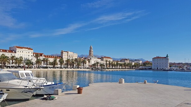 Un paseo marítimo con barcos amarrados, una torre destacada y una hilera de edificios a lo largo de la orilla.