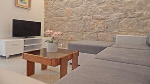 Una sala de estar con una pared de piedra, un televisor de pantalla plana, una mesa de centro de madera, un sofá gris y una pequeña planta en maceta.