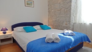 Una cama impecablemente tendida con ropa de cama azul y blanca, dos toallas enrolladas, una mesita de noche con lámpara y una pared de piedra.