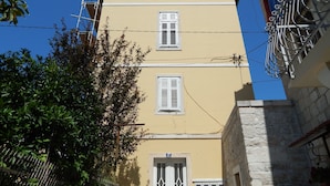 Un edificio de varios pisos con tres ventanas y una puerta, rodeado de follaje y una cerca metálica.