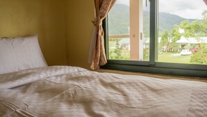 Ein Bett mit karierter Bettdecke und Kopfkissen, ein Fenster mit Vorhang sowie ein Blick auf Grünflächen und Gebäude.