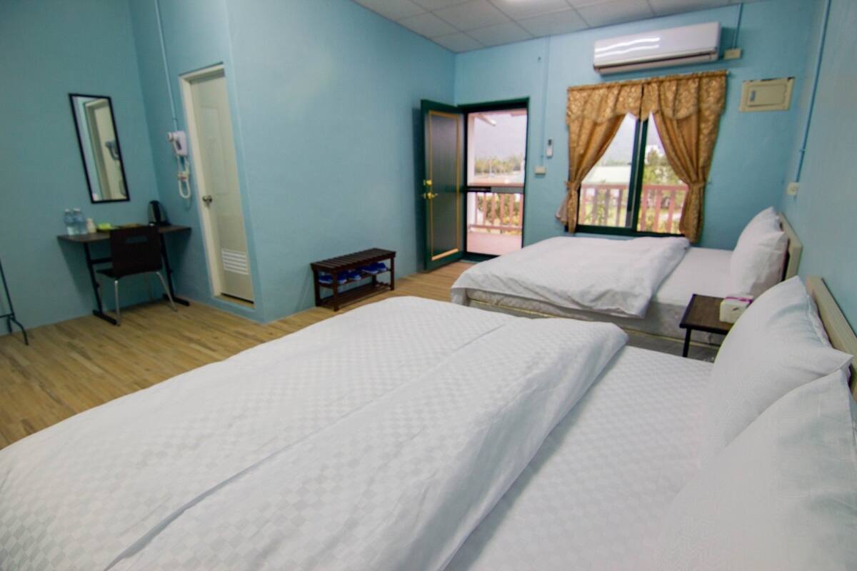 A hotel room with two beds, a desk, a chair, a small table, and a sliding door to a balcony.
