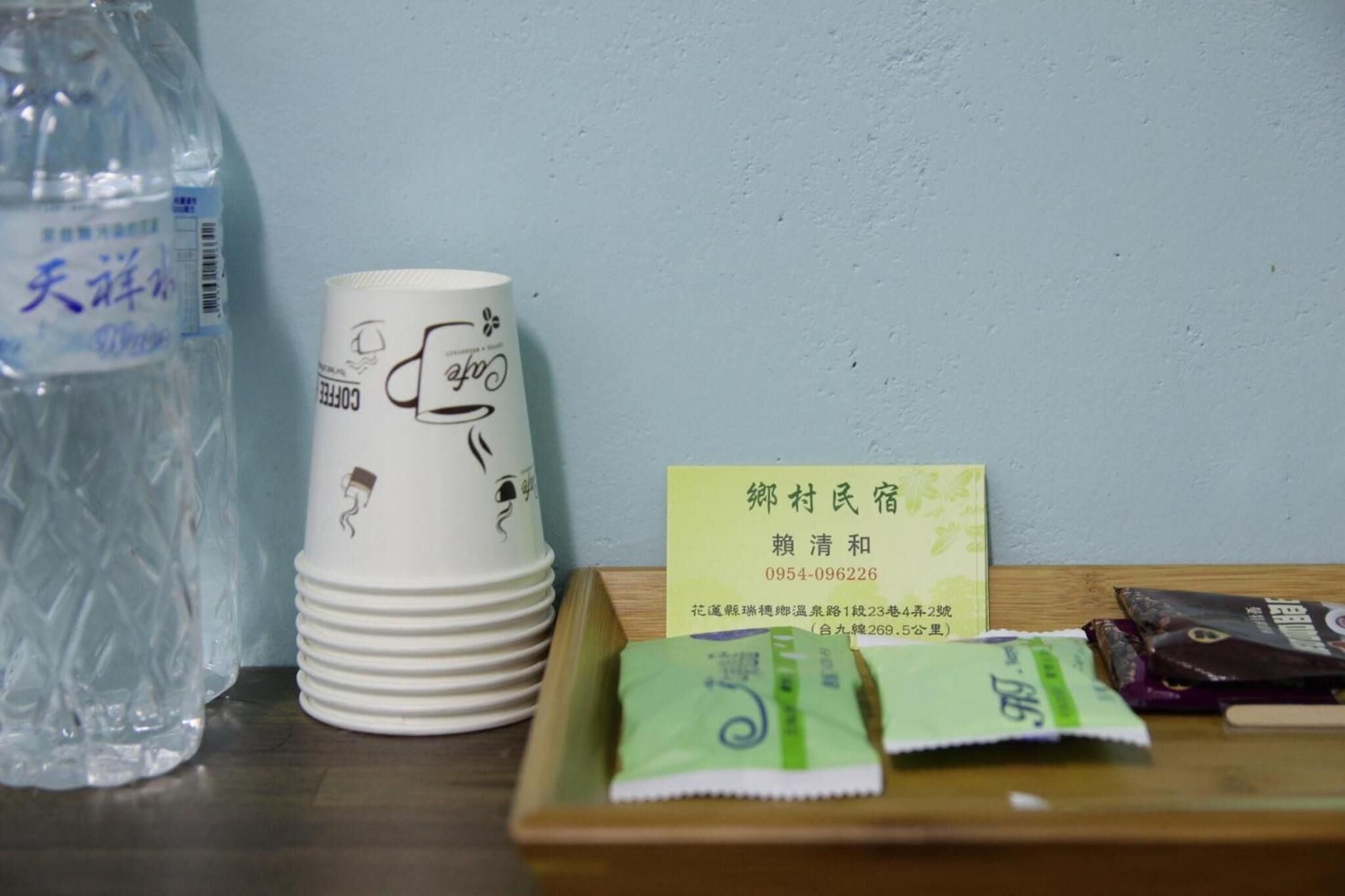 A stack of white paper cups with a coffee cup design, a plastic water bottle, and a wooden tray with tea packets and a business card.