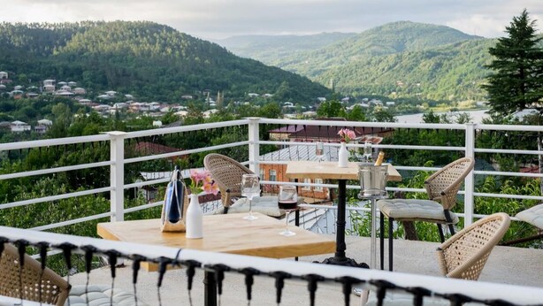 A terrace with wicker chairs and tables, overlooking a mountainous landscape with a village below. - Green House (Kutaisi)