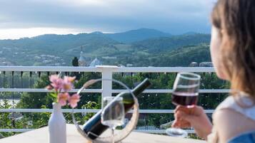 Una persona sentada a una mesa con una botella de vino y una copa, con vistas a un paisaje escénico de colinas y una iglesia.