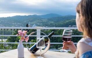 Una persona sentada a una mesa con una botella de vino y una copa, con vistas a un paisaje escénico de colinas y una iglesia.