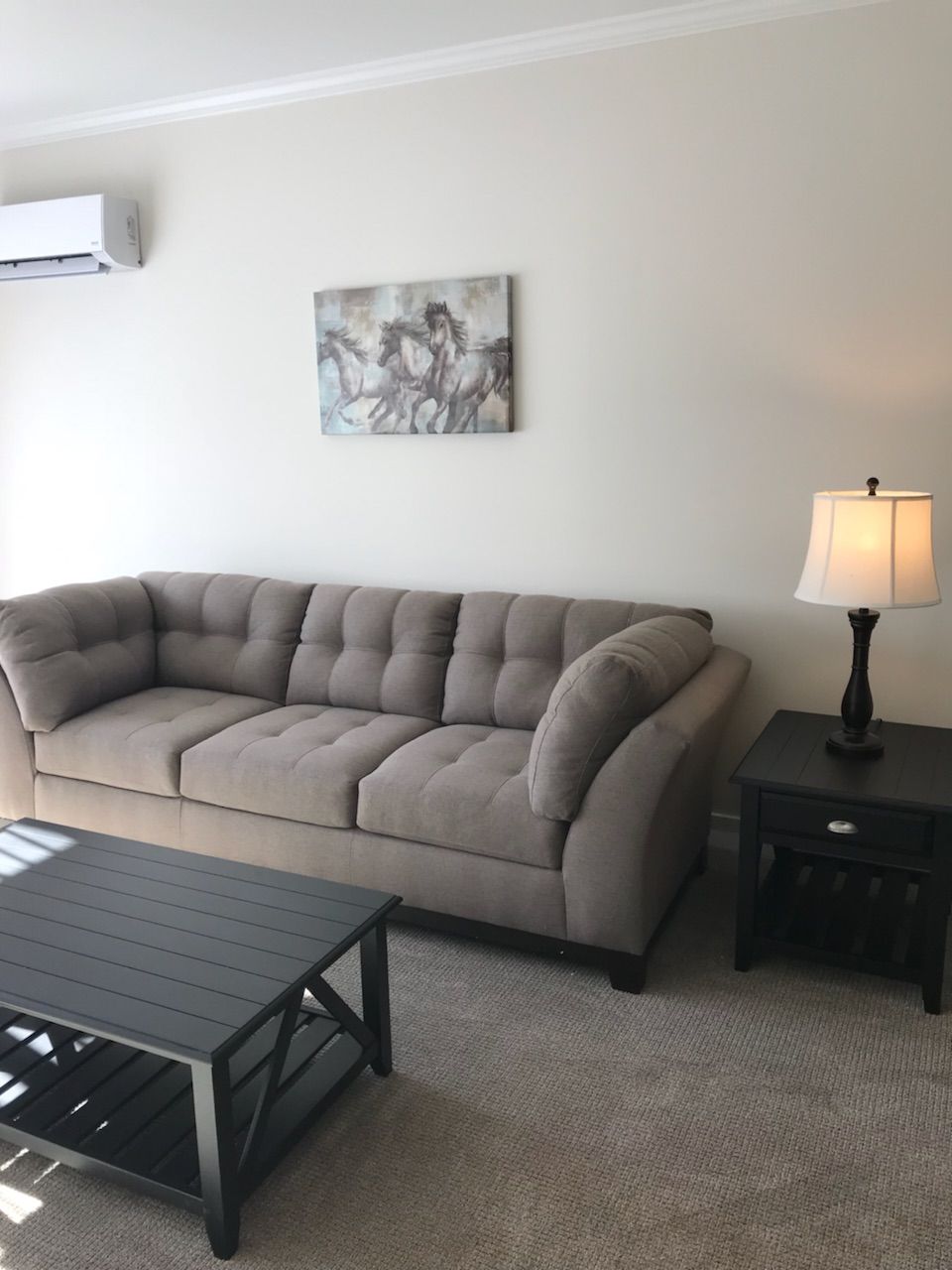 A living room with a sectional sofa, a small table, a lamp, and a wall-mounted air conditioner.