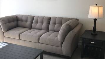 A living room with a sectional sofa, a small table, a lamp, and a wall-mounted air conditioner.