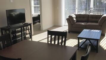 A living room with a TV, a sofa, a dining table, and chairs. There is a window with blinds and a door leading to another room.