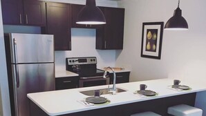 A modern kitchen with dark cabinetry, a stainless steel refrigerator, a stove, and a large island with bar stools. - The Hamlet (Saratoga Springs)