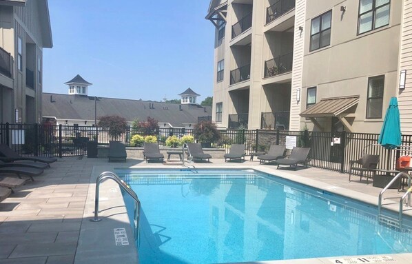 A swimming pool with lounge chairs and a fenced area, surrounded by multi-story buildings with balconies. - The Hamlet (Saratoga Springs)