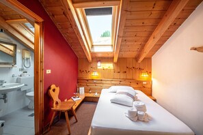 Una stanza con un lucernario, travi a vista in legno, un letto con asciugamani, una sedia di legno, un comodino, una lampada e un bagno con lavandino e WC.