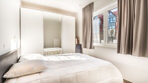 Un dormitorio con cama, armario, ventana con cortinas y vistas a edificios.