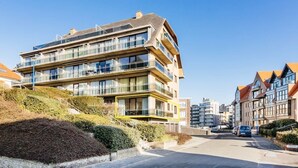 Un edificio residencial de varios pisos con balcones, cielo despejado y una calle en primer plano.