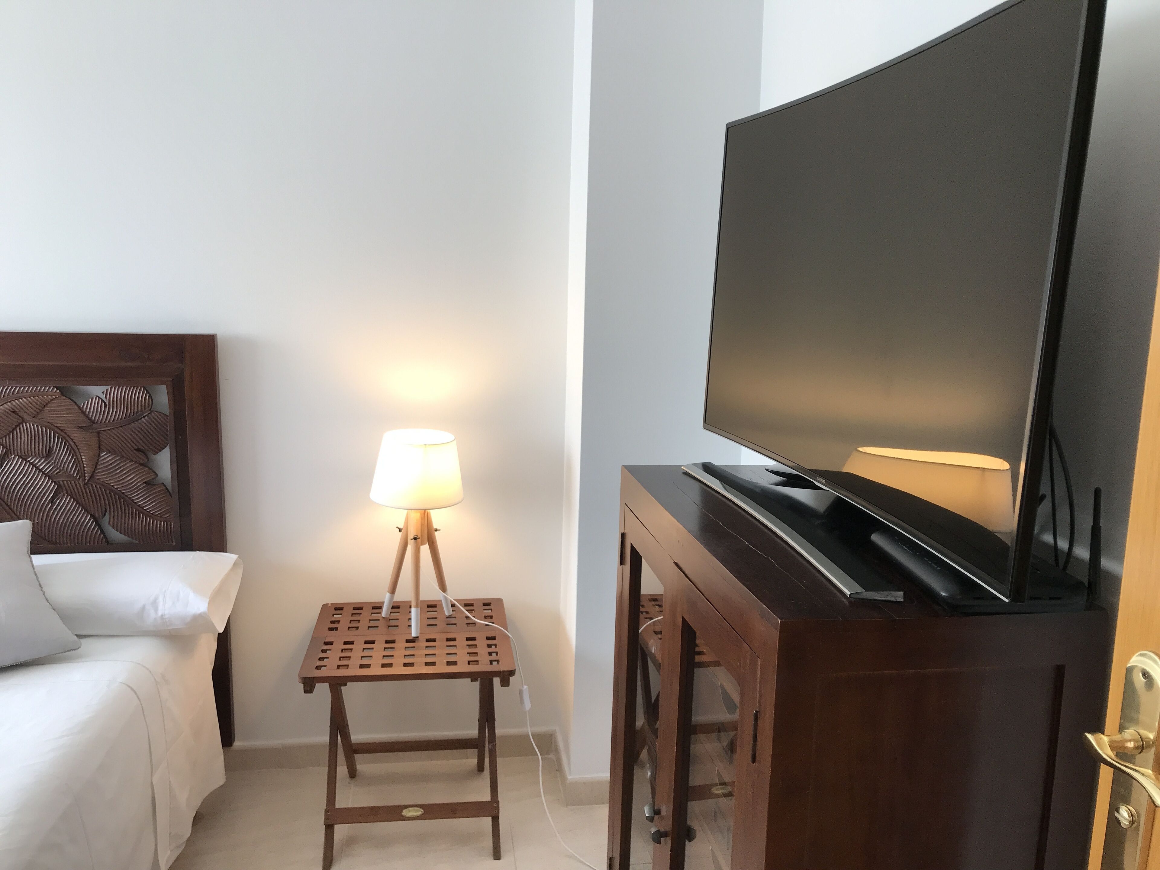 Une chambre d’hôtel avec un lit, un téléviseur posé sur un meuble en bois, une lampe sur une petite table et une tête de lit en bois.