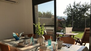 Comedor con una mesa preparada para cuatro, incluyendo platos, tazones, vasos y cubiertos. Un jarrón con una planta está sobre la mesa, y se ve un jardín y una valla desde el exterior.