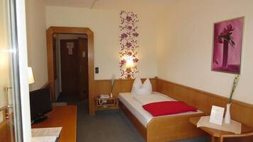 Habitación de hotel con cama, escritorio, silla, televisor, lámpara, una obra de arte en la pared y una puerta.