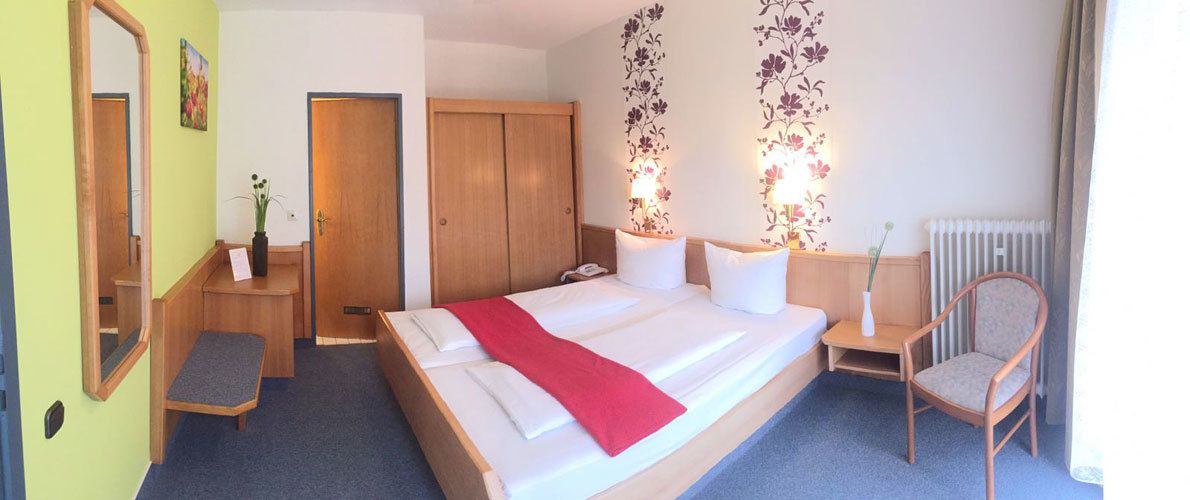 A hotel room with a large bed, a wooden wardrobe, a bedside table with a vase, and a chair.