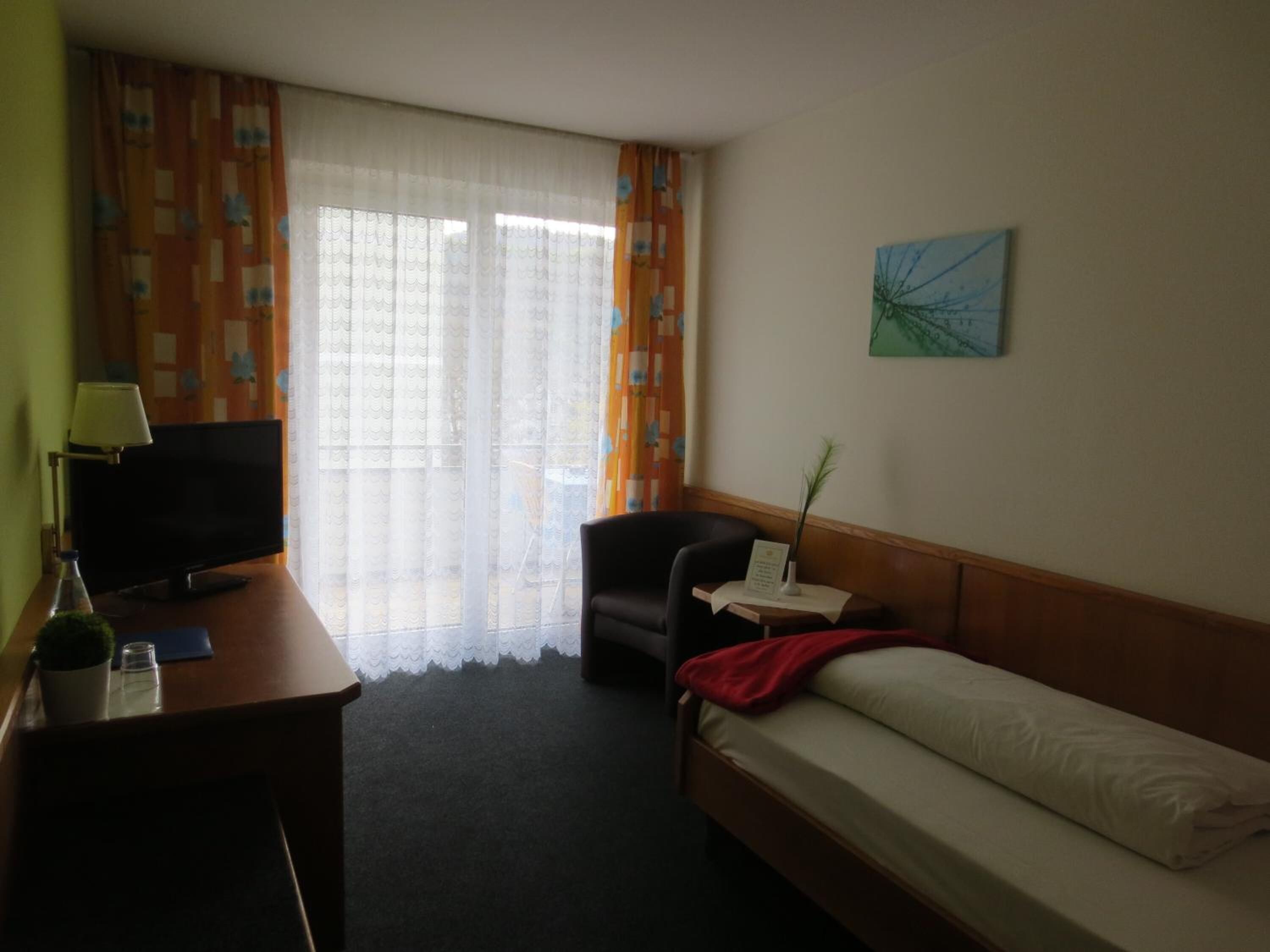 Ein Hotelzimmer mit einem Bett, einem Schreibtisch mit Fernseher, einem Sessel und einem kleinen Tisch mit Pflanze.