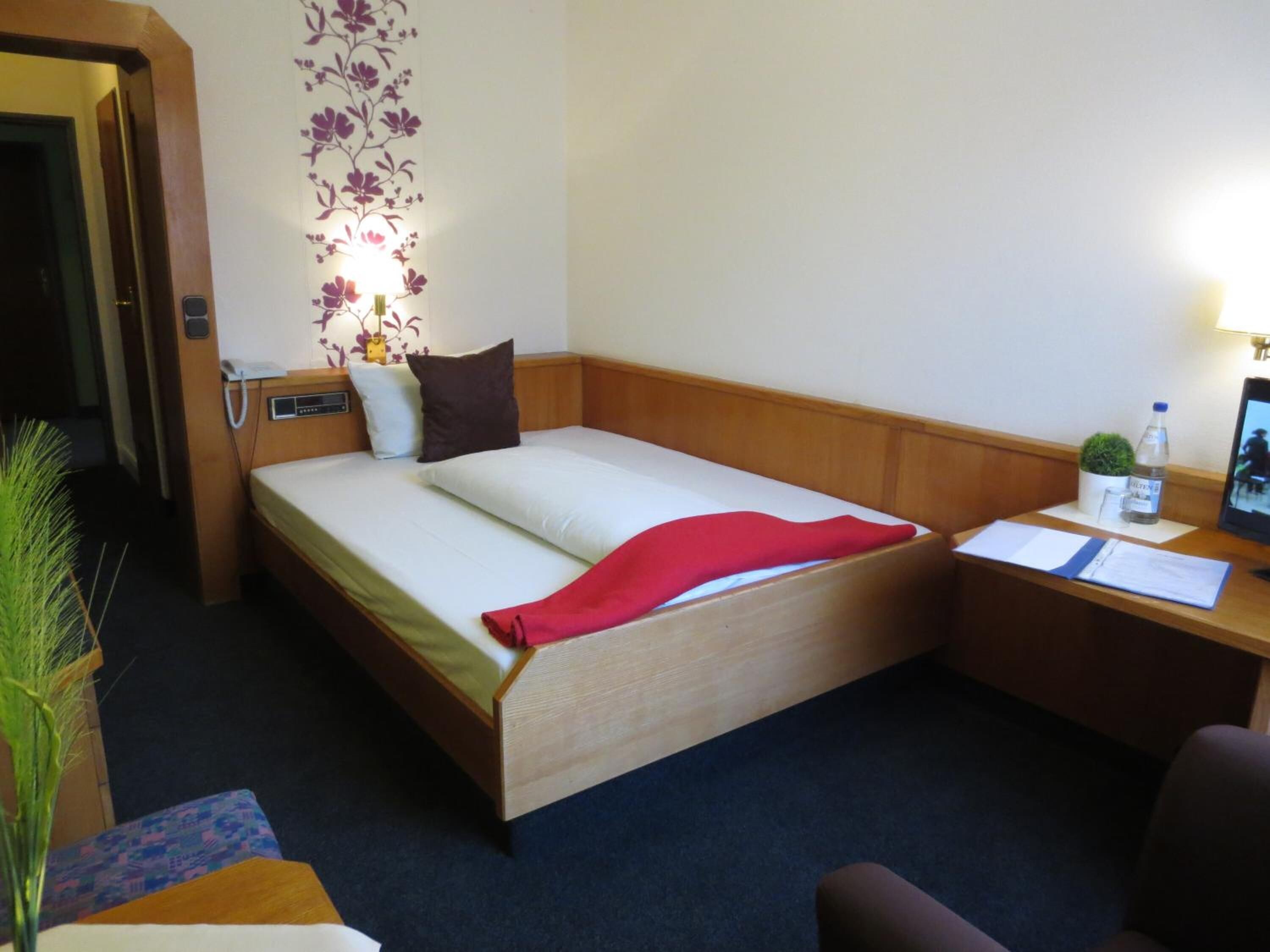 A hotel room with a bed, bedside table, lamp, and a small potted plant.