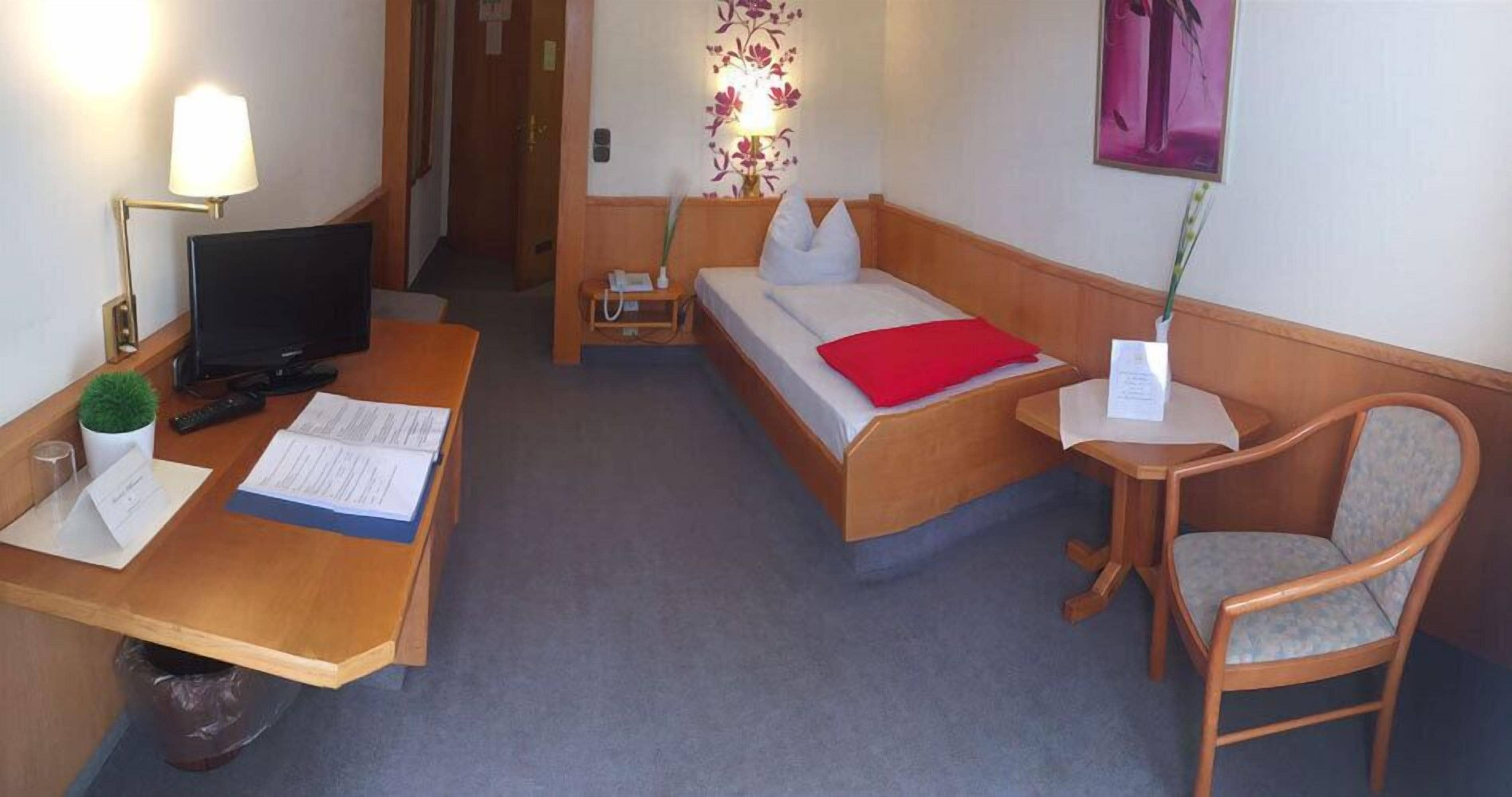 A hotel room with a desk, two beds, a chair, a TV, and a small table with a lamp.