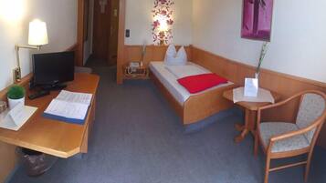 A hotel room with a desk, two beds, a chair, a TV, and a small table with a lamp.