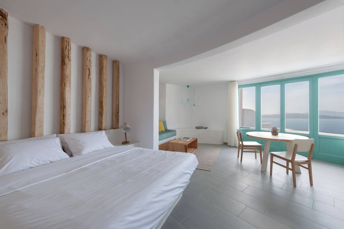Habitación de hotel moderna con una cama grande, cabecero de madera, una mesa redonda con sillas y vistas amplias a través de grandes ventanales.